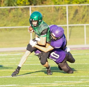 Two football players collide during a tackle, one in green and one in purple uniforms.