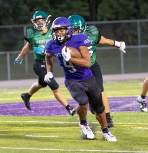 High school football player in purple uniform carries ball while running downfield during night game.