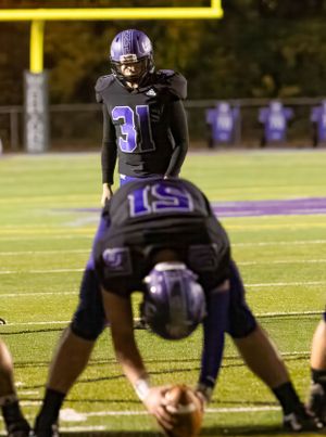 Football players in purple uniforms line up before snap on illuminated field at night.