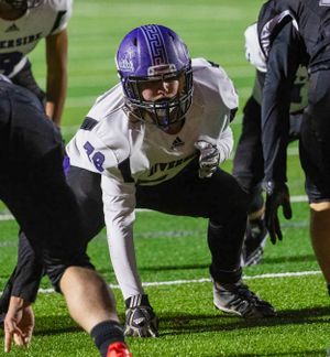 Football player in purple uniform and helmet crouches in three-point stance.