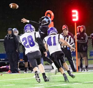 Players leap for football pass during nighttime game with scoreboard visible.