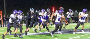 High school football players in purple and white uniforms engage in a running play during a nighttime game.