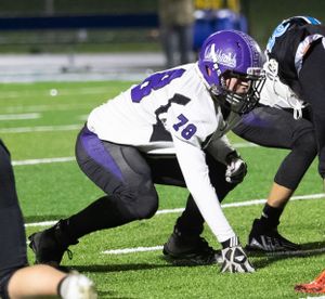 Football lineman in purple and white uniform takes stance at line of scrimmage during game.