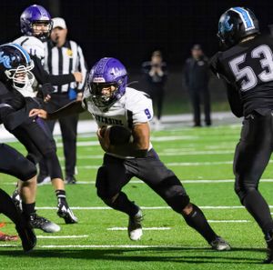 Running back in purple and white uniform dodges defenders during nighttime football game.