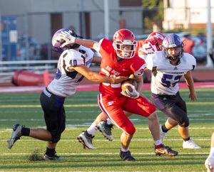 Football player in red uniform carries ball between defenders during daytime game.