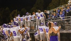 Football team in purple and white uniforms celebrates along sideline bleachers at night.