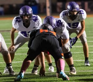 Football players in purple and white uniforms line up against opponent in orange and black at the line of scrimmage.