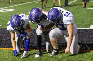 Three football players in purple helmets huddle together on the sideline during a game.