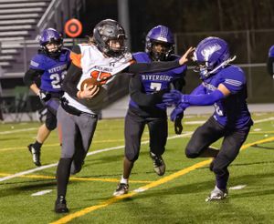 Football player in black jersey carrying ball while being chased by defensive players in purple uniforms on field at night.