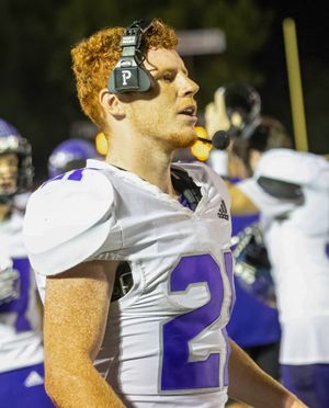 Football player wearing white uniform and headphones stands on sideline during night game.