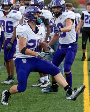 Football players in purple and white uniforms huddle on sideline during game.