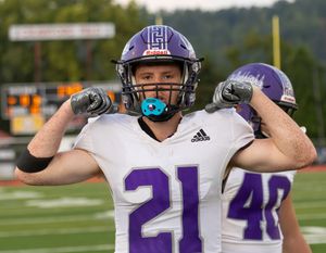 Football player in white jersey number 21 with purple helmet flexes on field.