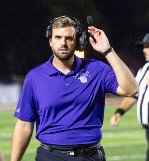 Football coach in purple polo shirt wearing headset communicates from sideline during night game.