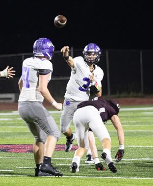 High school quarterback throws football during nighttime game while teammate protects from defenders.