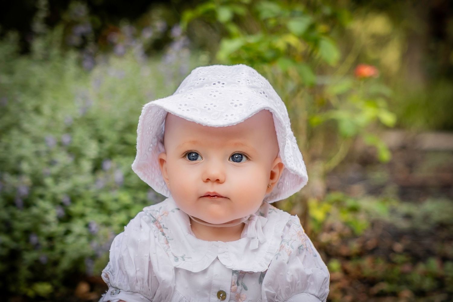 Adorable baby in white bonnet and dress sitting outdoors in a lush garden.