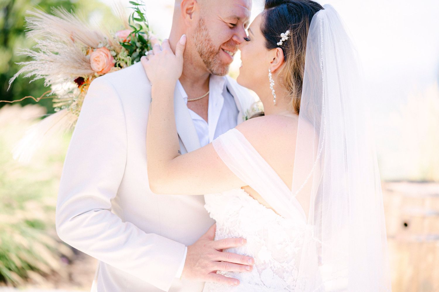 A romantic wedding couple embraces outdoors in soft sunlight with a flowing white veil and tropical greenery backdrop.