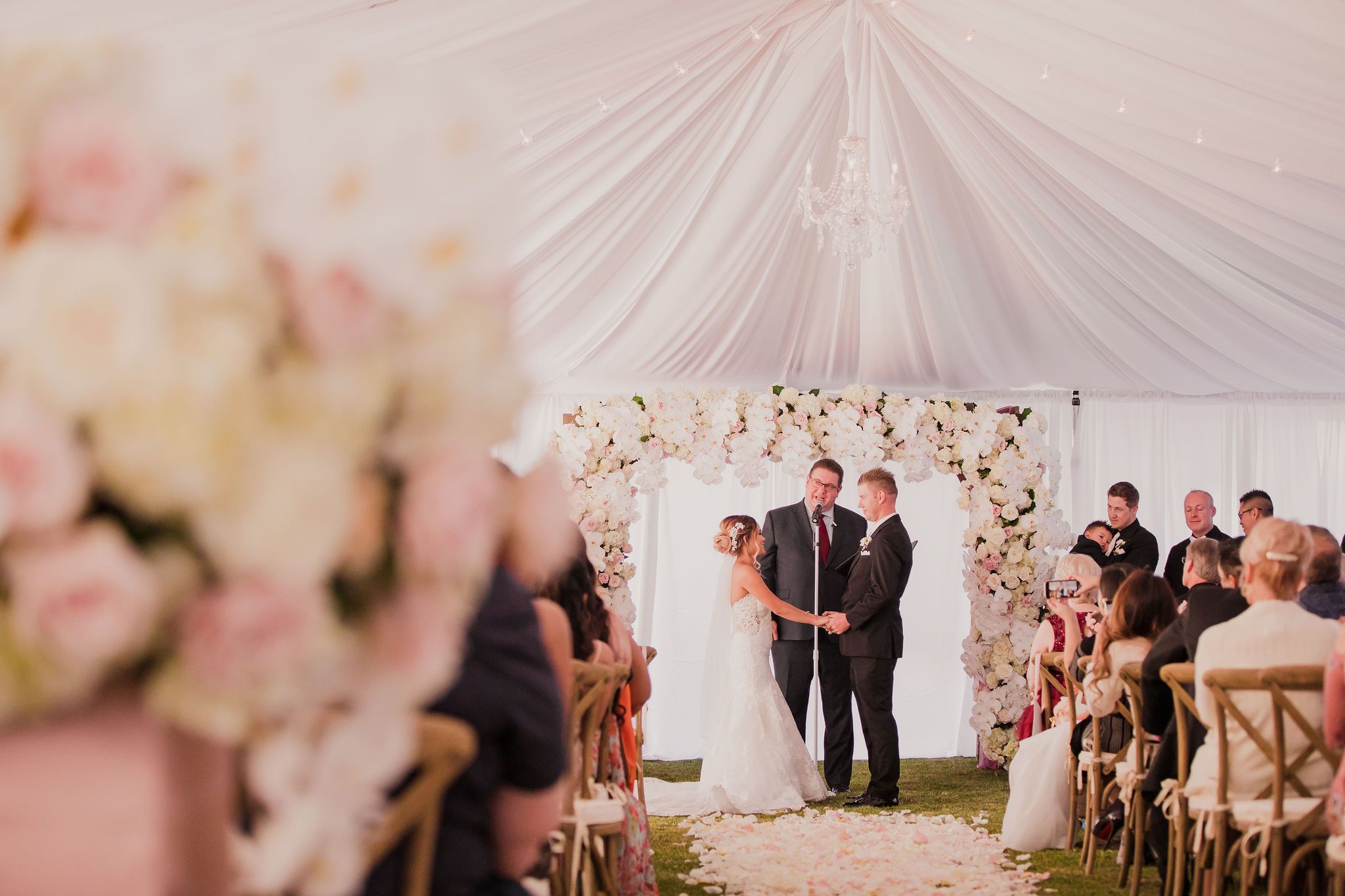 A wedding ceremony takes place under a white tent with pink floral decorations and guests seated in rows.