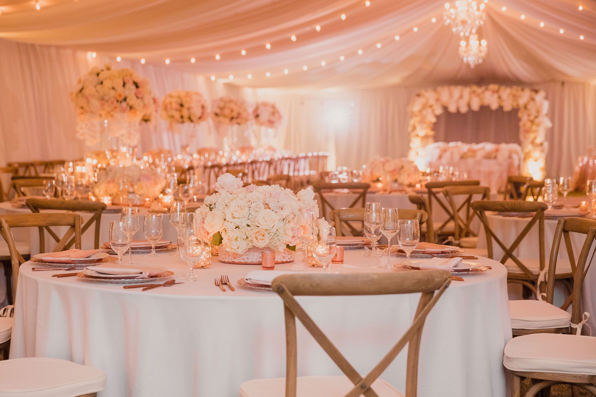 Elegant wedding reception venue with white draping, crystal chandeliers, and romantic warm lighting illuminating round tables.