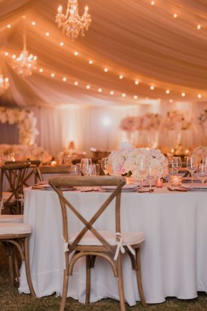 Elegant wedding reception venue with round tables, white linens, wooden cross-back chairs, and warm twinkle lights overhead.