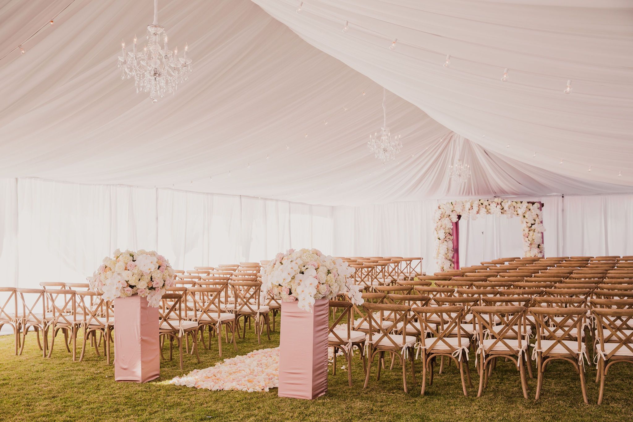 Elegant outdoor wedding ceremony setup with pink pillars, wooden crossback chairs and draped white tent ceiling with chandelier.