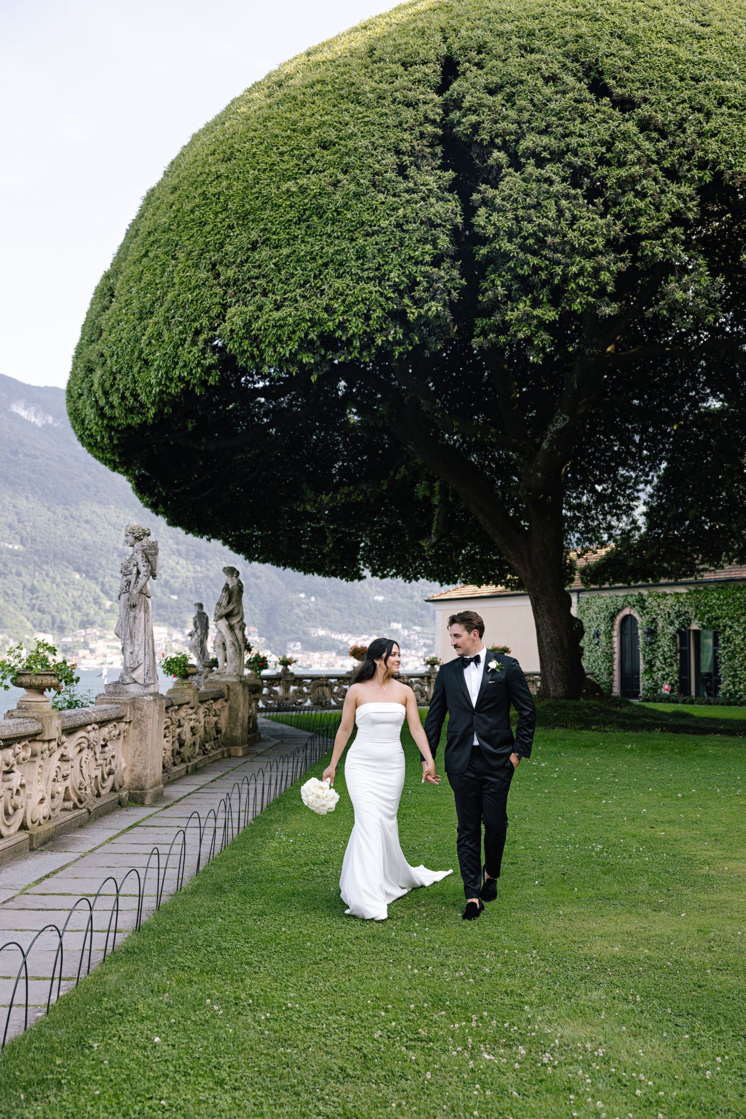 A couple walks down an outdoor aisle lined with flowers and cypress trees leading to an elegant villa entrance.