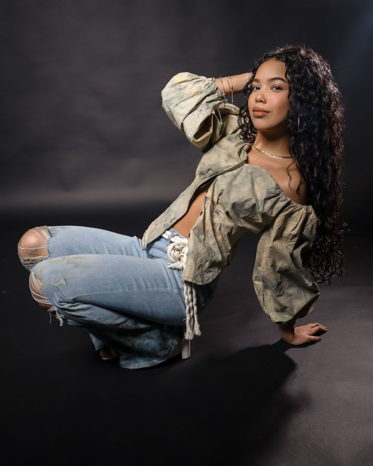 A person in a camouflage jacket and ripped jeans poses dramatically against a dark background for a senior picture in studio Charleston, SC