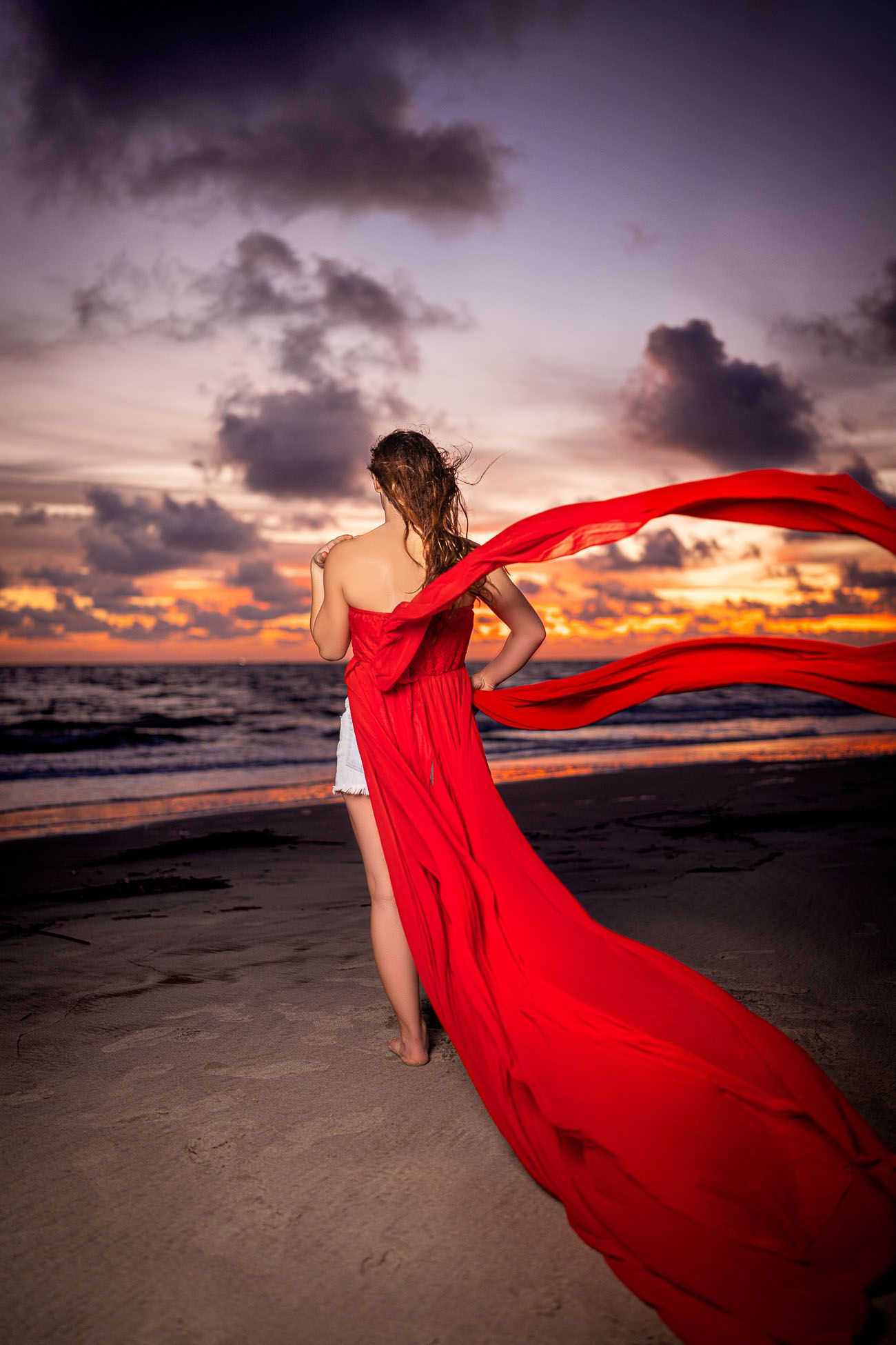 High school senior portrait on Folly Beach at sunset with flowing red dress and ocean waves in Charleston SC
