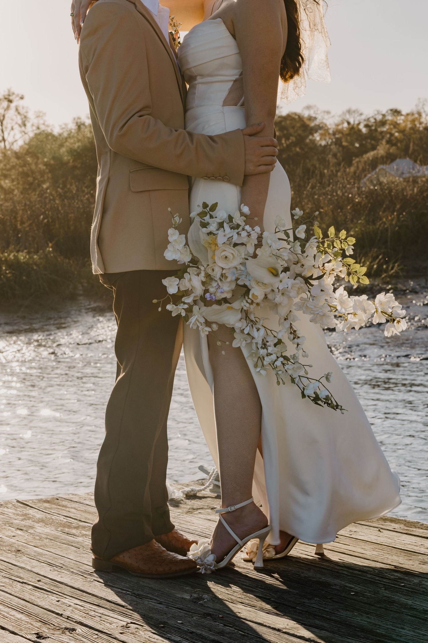 Bride and groom embracing on a wooden dock during golden hour with a white floral bouquet by the water.
