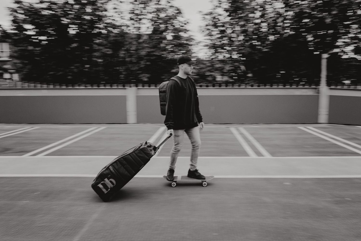 A person in casual attire pulls a rolling suitcase across an empty parking structure in black and white.
