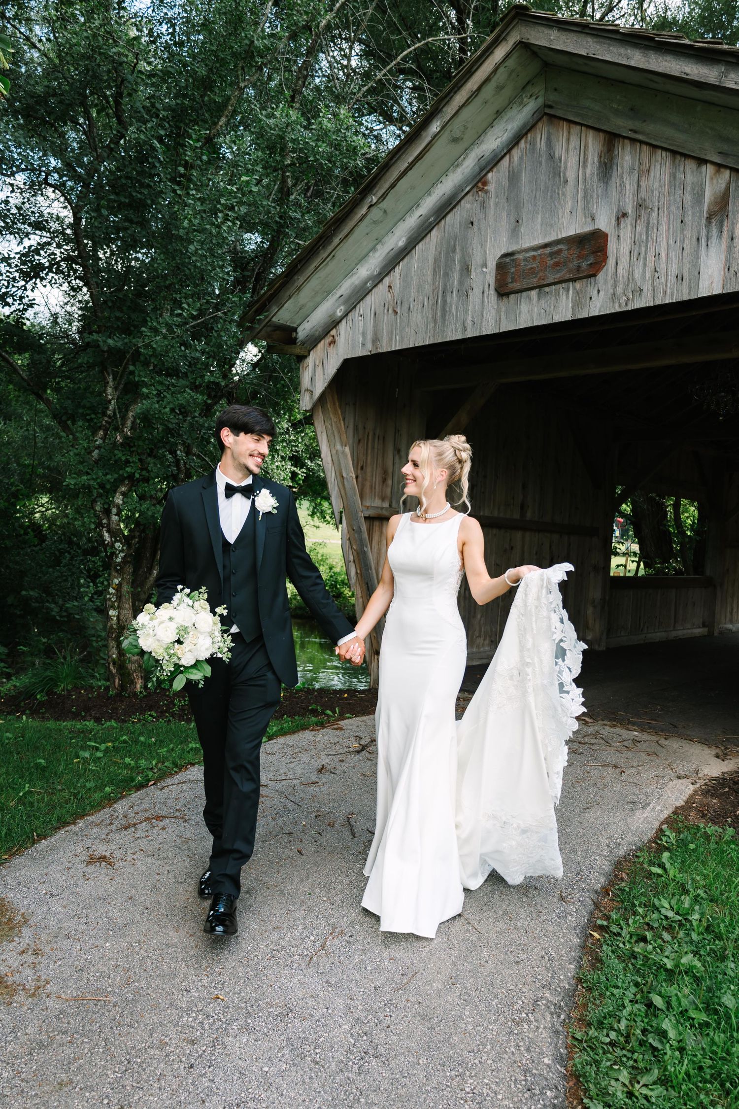 A couple in formal wedding attire walks along a gravel path beside a rustic wooden covered bridge.