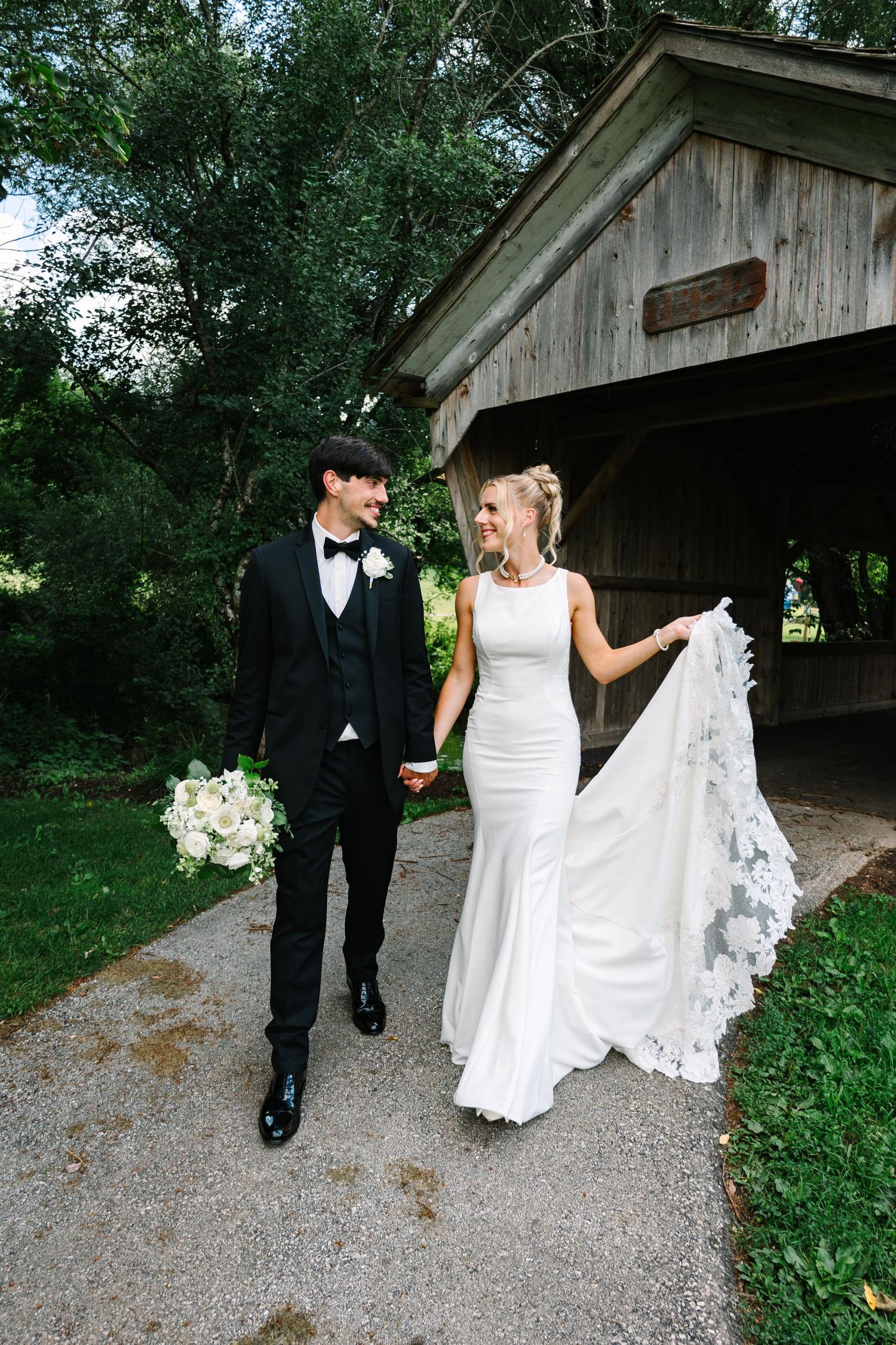 A couple in formal wedding attire walks together on a gravel path near a rustic wooden covered bridge.