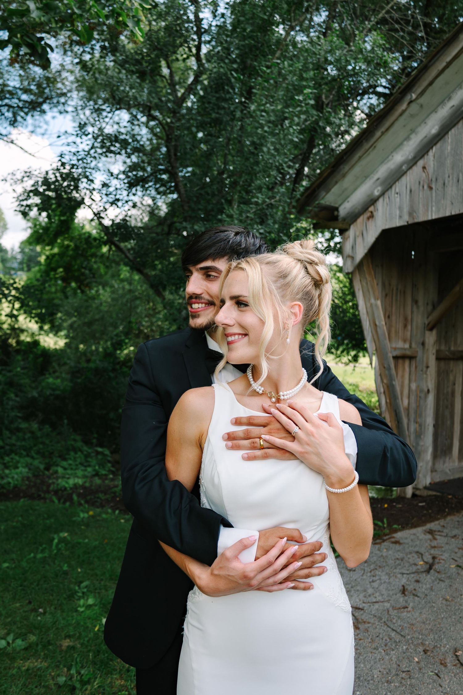 A romantic embrace captured in a rustic outdoor setting with a weathered barn and lush greenery in the background.