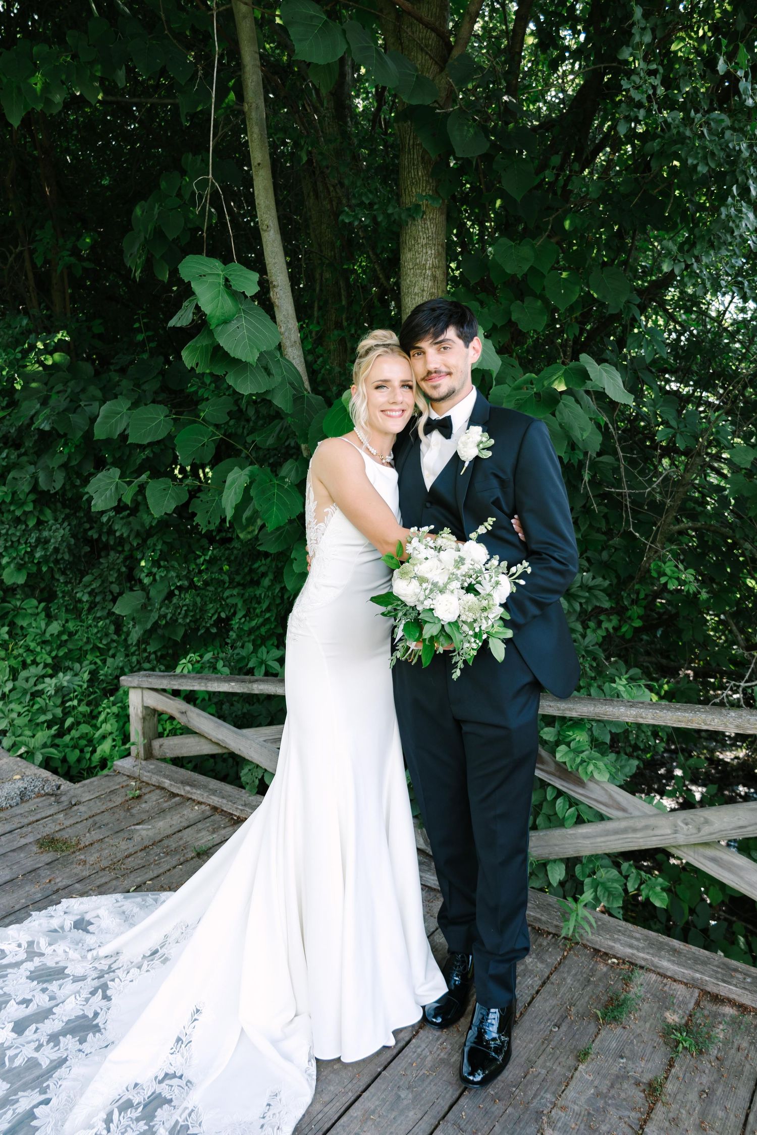 A couple in wedding attire poses together on a wooden deck surrounded by lush green foliage.