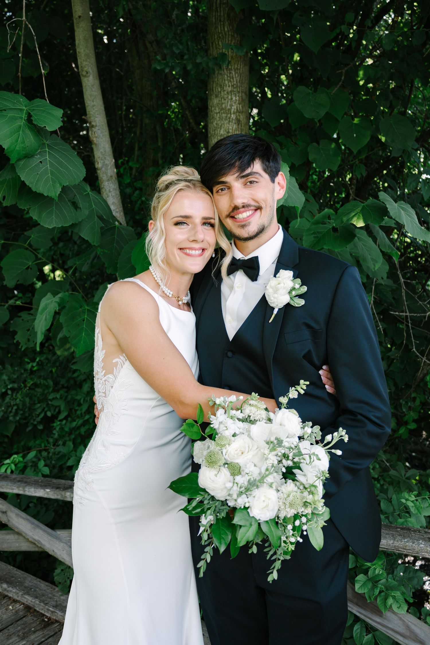 A newlywed couple poses with white floral bouquet against lush green foliage background.