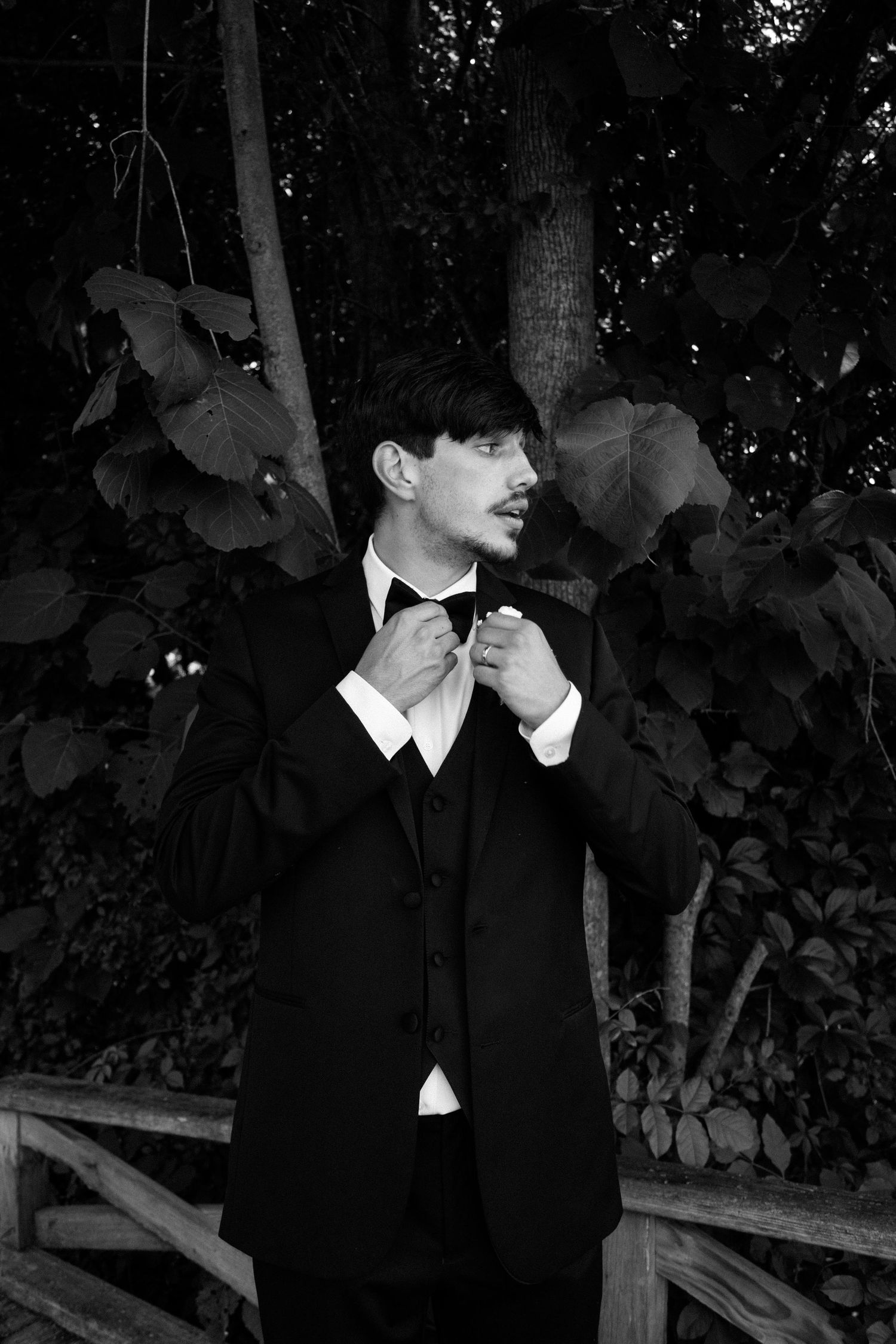 A black and white artistic portrait shows a figure in formal attire against a dark leafy background.