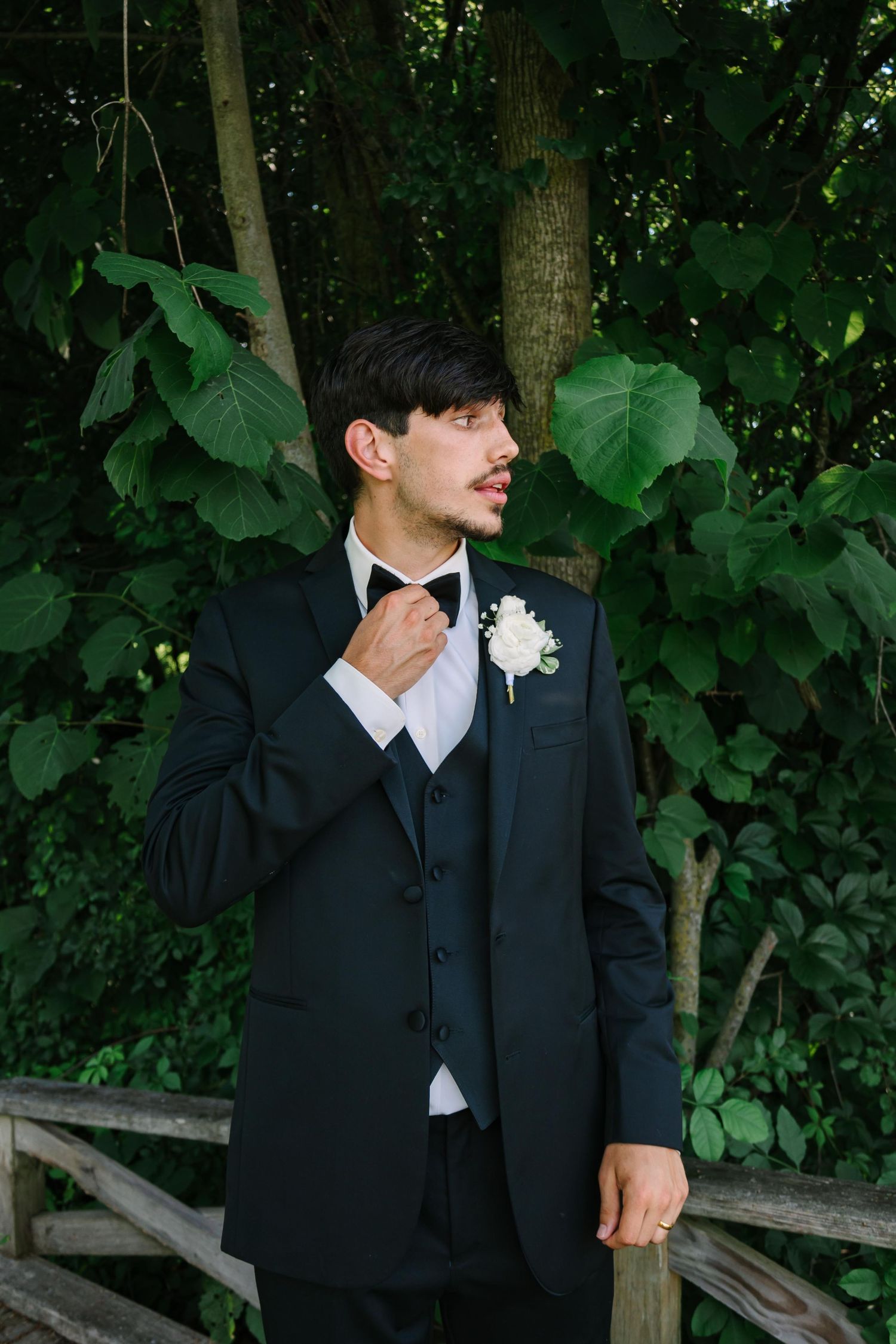 A groom in a black suit stands on a wooden deck with trees and greenery in the background.