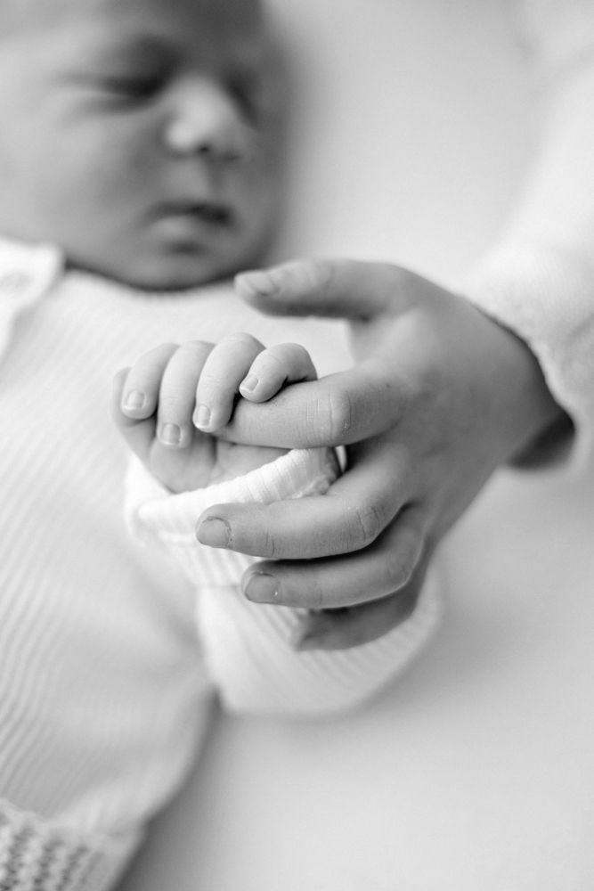 A black and white image of a newborn's tiny hand grasping their siblings finger.