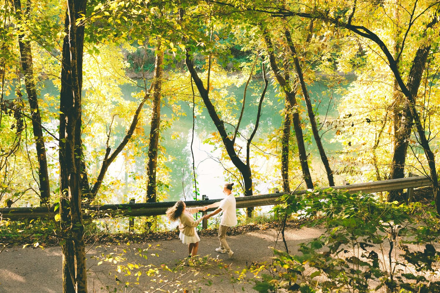 Two people walk along a scenic autumn path with golden yellow leaves and reflective water views in the background.