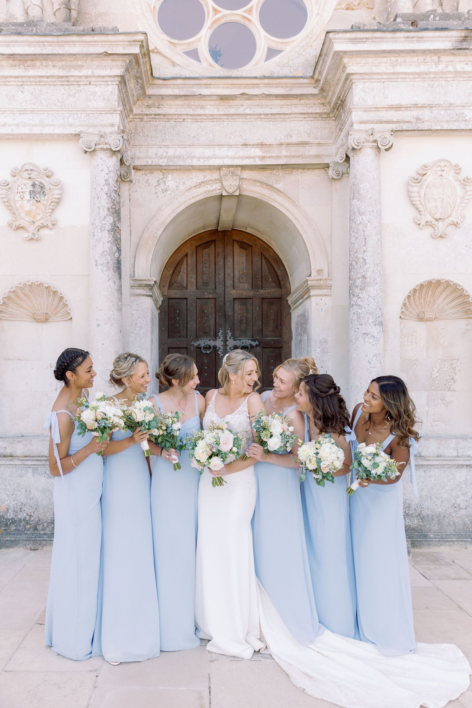 A bridal party in light blue dresses and white gown pose together against a historic stone church facade.