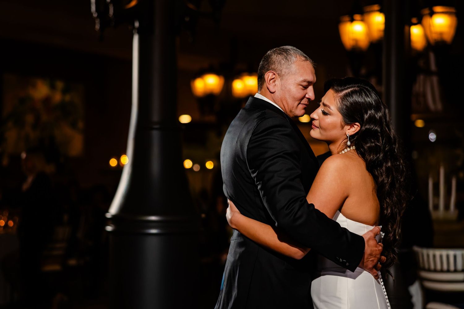 A romantic couple shares an intimate dance moment under warm glowing lights at an evening celebration.