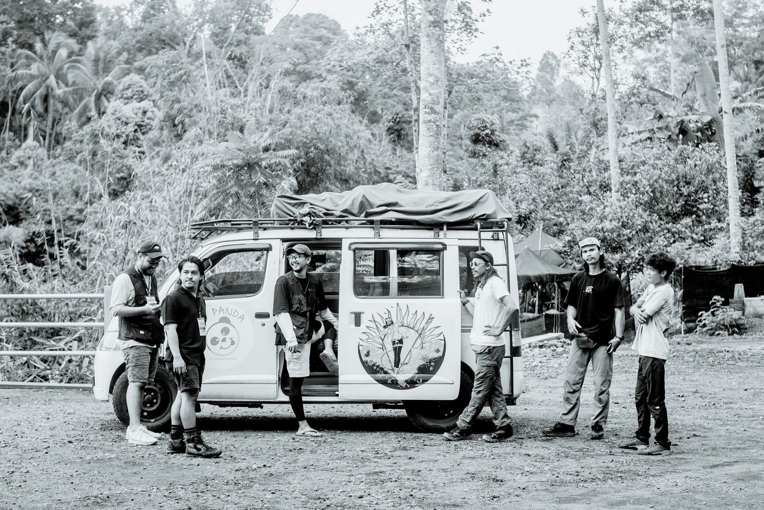 A vintage black and white photo shows a group gathered around a classic VW bus with surfboards on top in a forest setting.