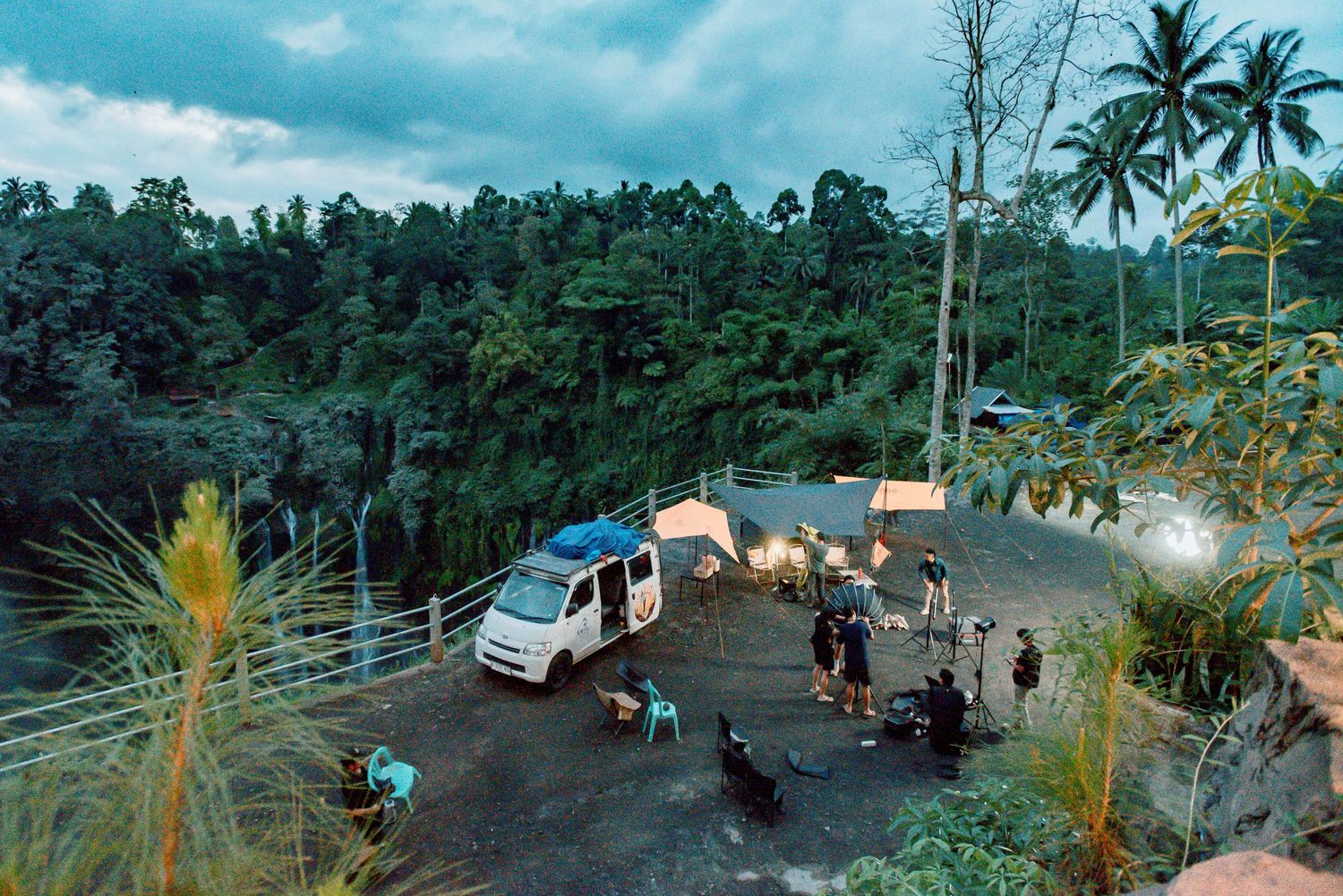 Vehicles and people gather at a scenic mountain viewpoint surrounded by palm trees and forest at dusk.