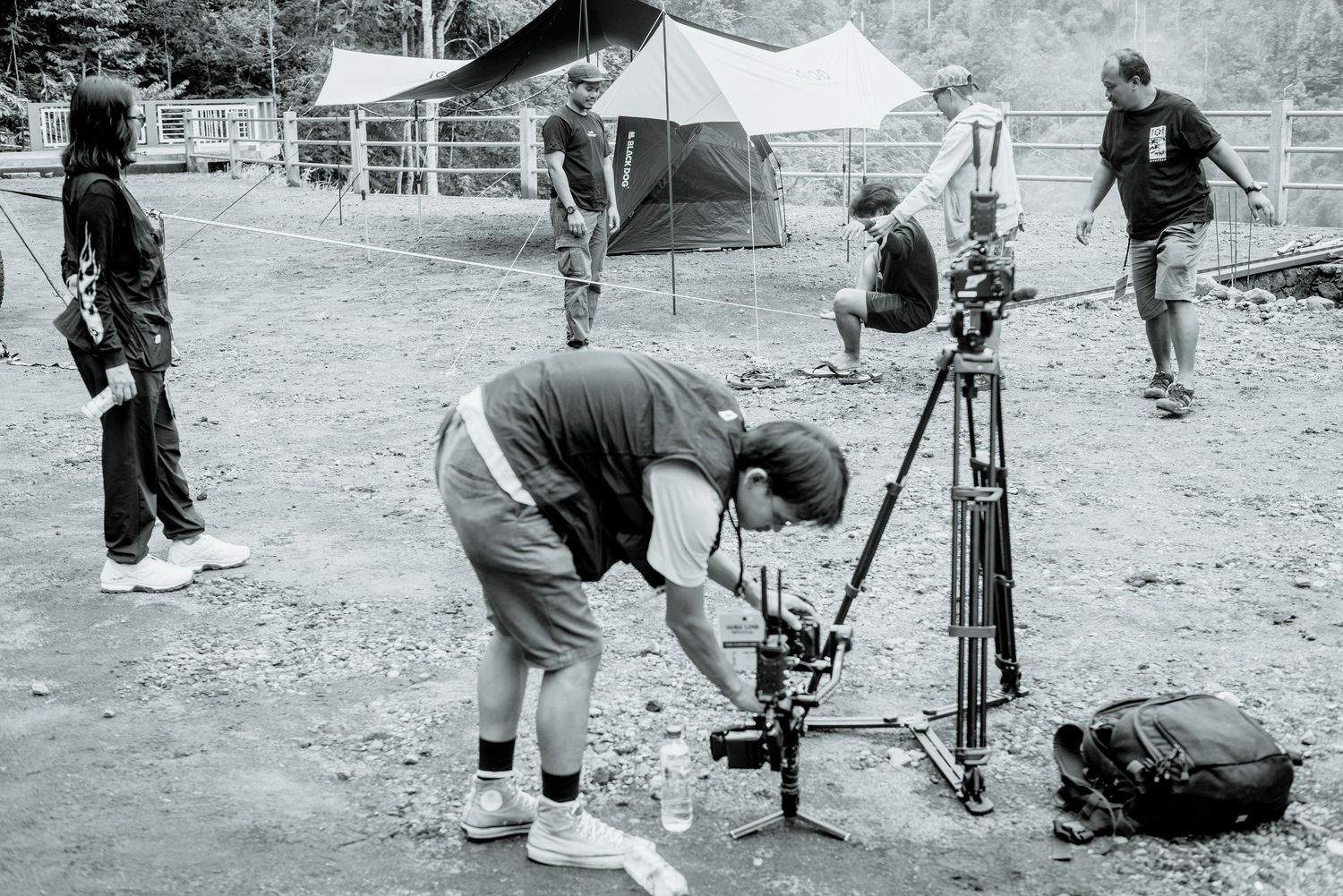 Behind the scenes black and white photo of a film crew setting up camera equipment on location at a campsite.