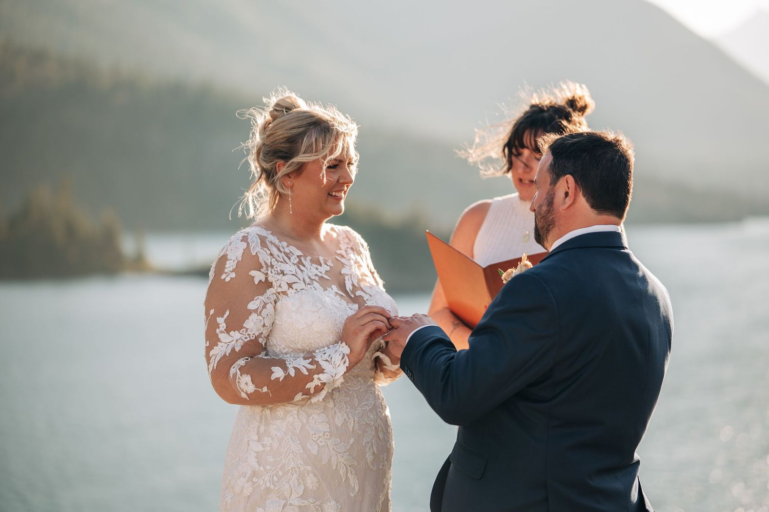A couple exchanges rings during their wedding ceremony by a scenic mountain lake at sunset.