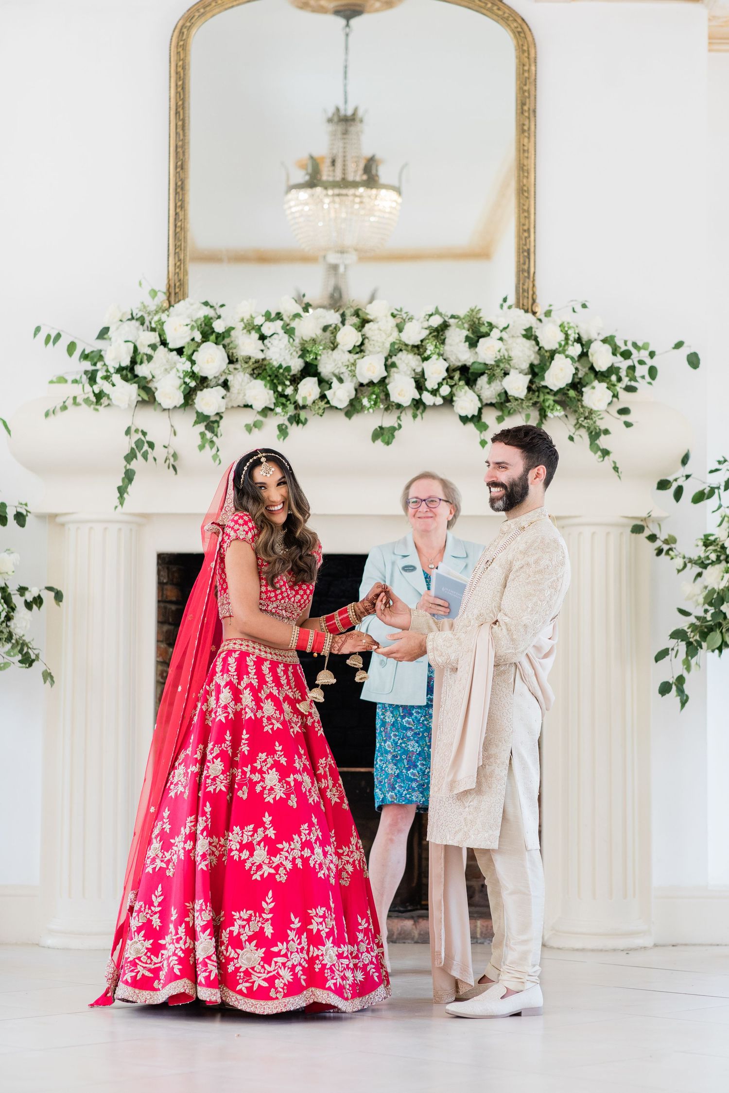 A bride in a red embroidered lehenga twirls in an elegant white room decorated with floral garlands and mirrors.