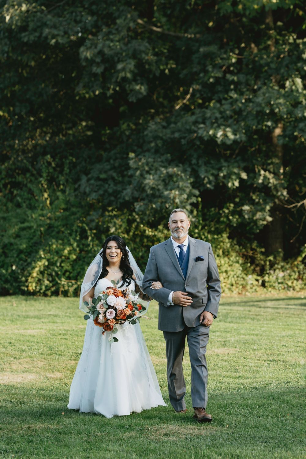 A couple in wedding attire walks together on a sunny lawn with green trees in the background.