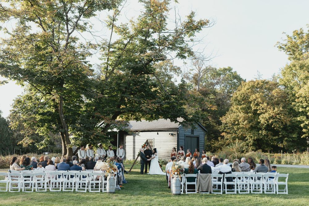 Panoramic view of an outdoor wedding ceremony setup with white chairs on a grassy lawn surrounded by trees.