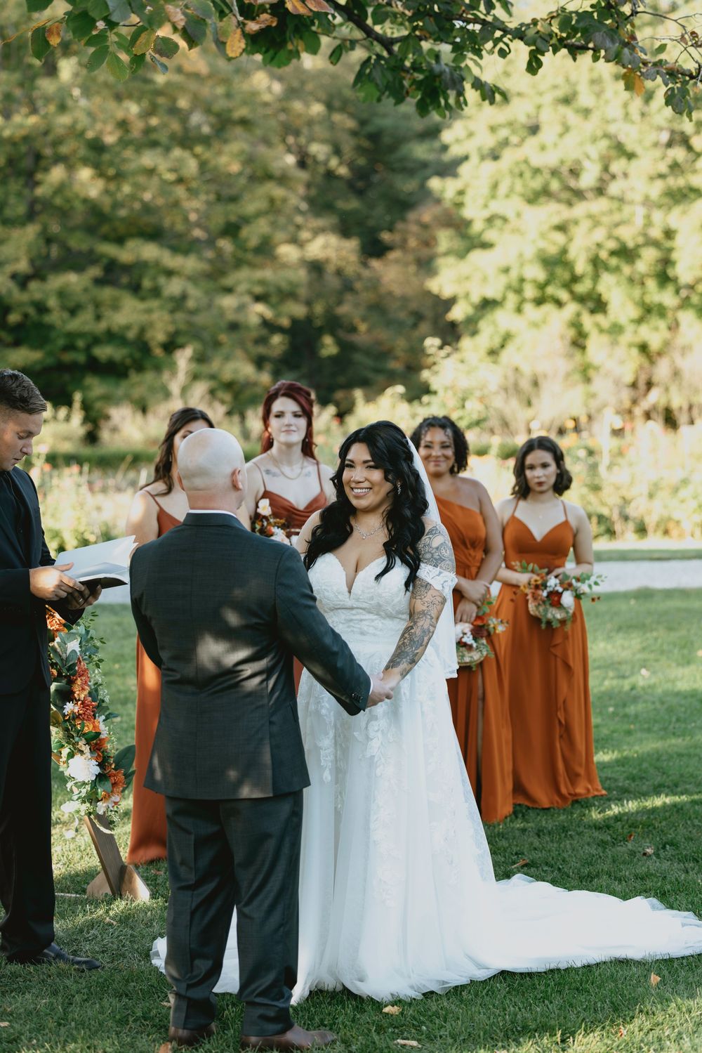 An outdoor wedding ceremony takes place on a sunny day with bridesmaids in burnt orange dresses standing in a row on grass.