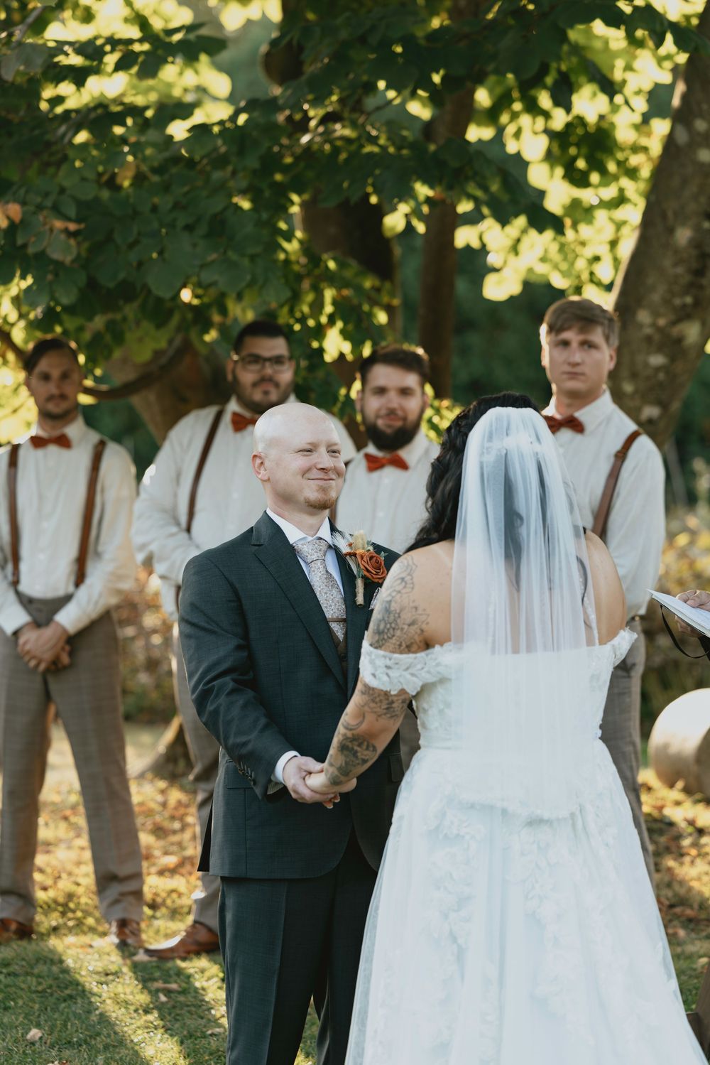 Wedding ceremony taking place outdoors under trees with sunlight filtering through green leaves.