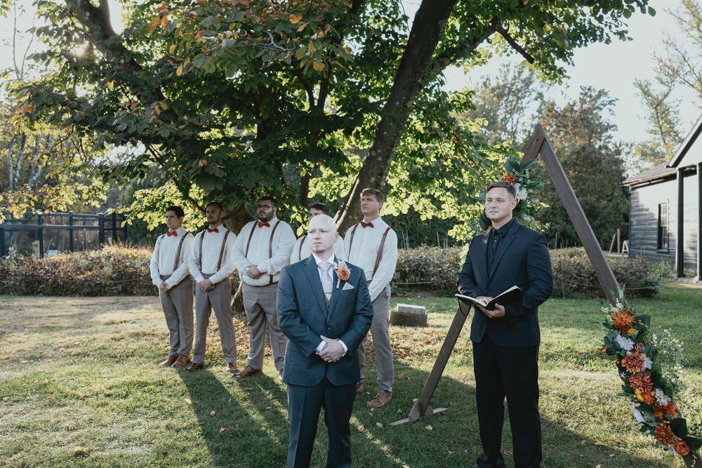 A wedding party stands outdoors in a garden setting with groomsmen in gray suits and officiant during a fall ceremony.